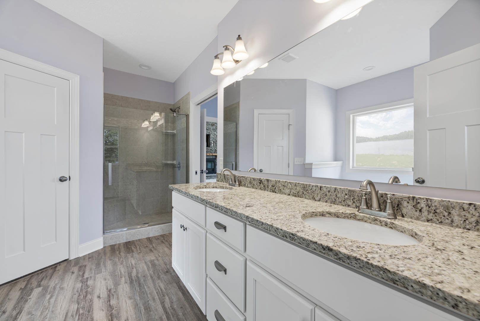 Bathroom featuring a wide framed mirror above a double sink vanity with white cabinetry, silver faucets, glass shower door, and a window overlooking a lake.