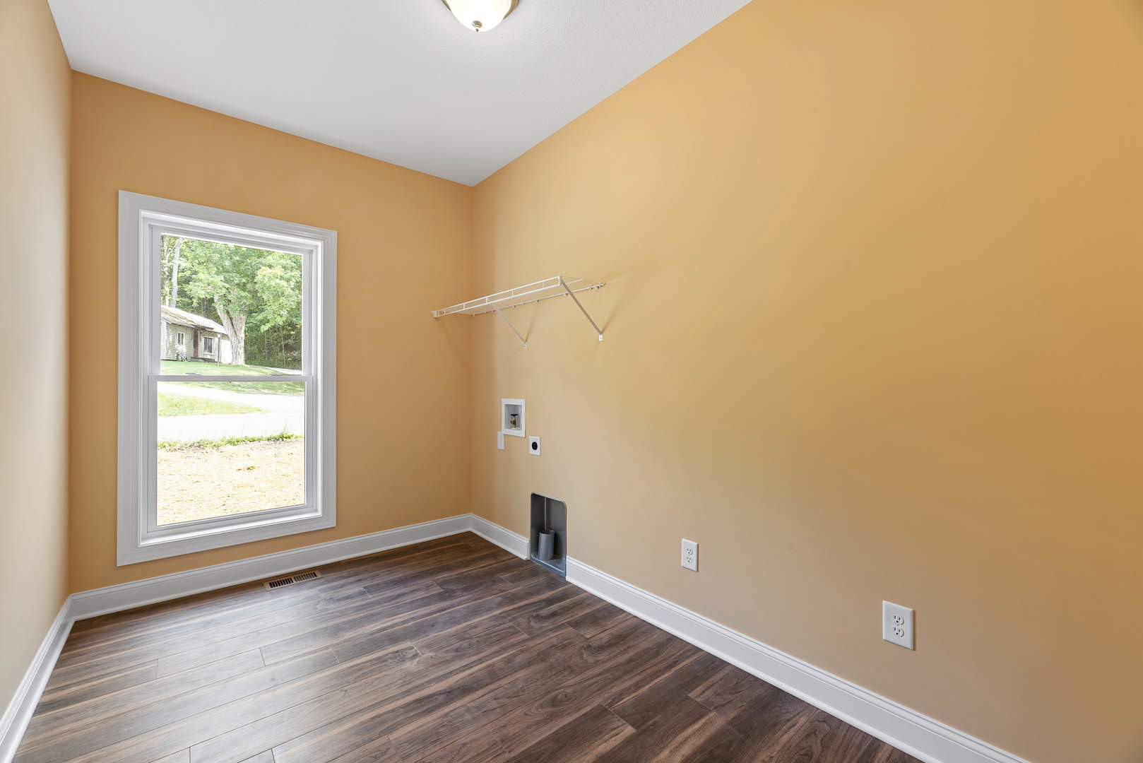Sunlit room with wide wood plank flooring, white baseboards, plaster walls, large window overlooking neighboring house, built-in white wall shelf, silver trash can, and visible
