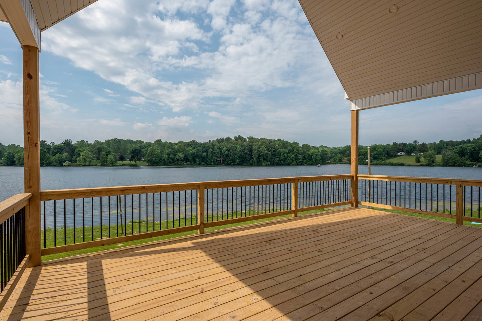 Wooden deck with metal railing, overlooking a calm lake surrounded by trees under a blue sky with scattered clouds