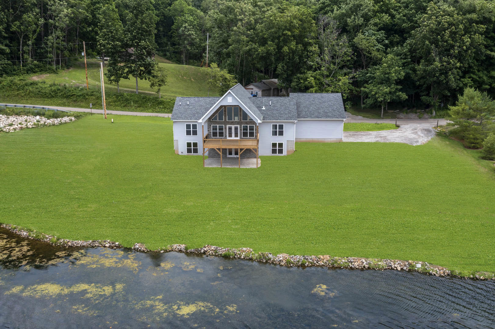 Two-story house with wood deck overlooking expansive green lawn, mature trees, and a pond bordered by white rocks