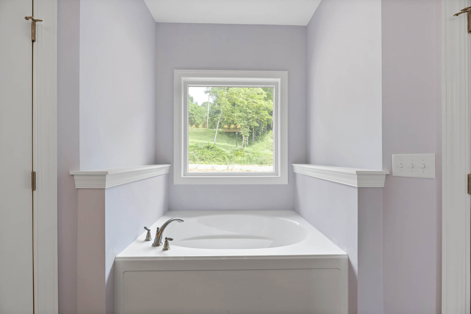 Freestanding white bathtub with silver faucet beneath large window overlooking forest, surrounded by light tile walls and four-switch panel
