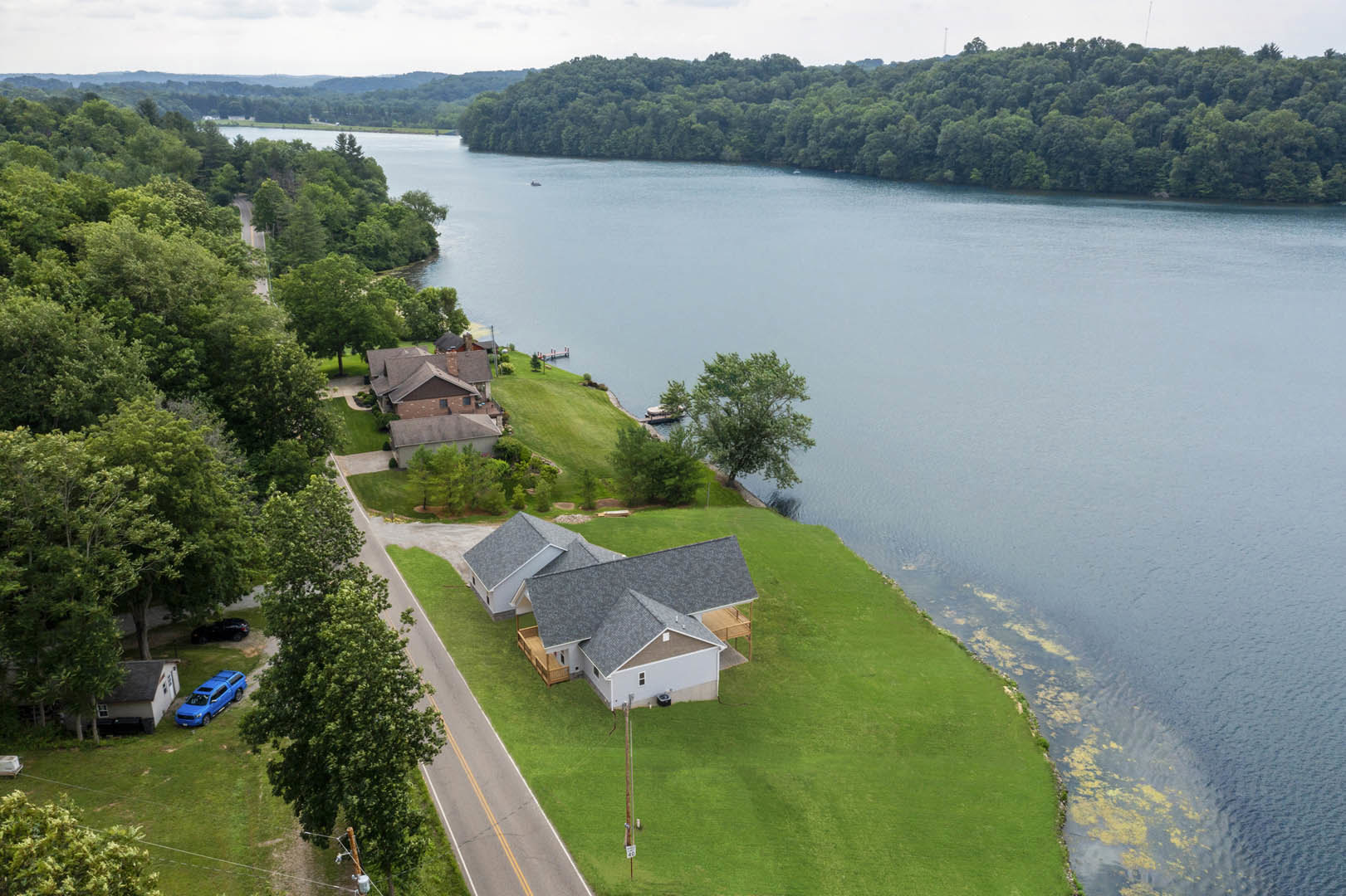 Two-story house with light siding and dark roof sits beside a river, surrounded by green lawn, mature trees, and a blue truck parked on grass; water and sky visible in background.