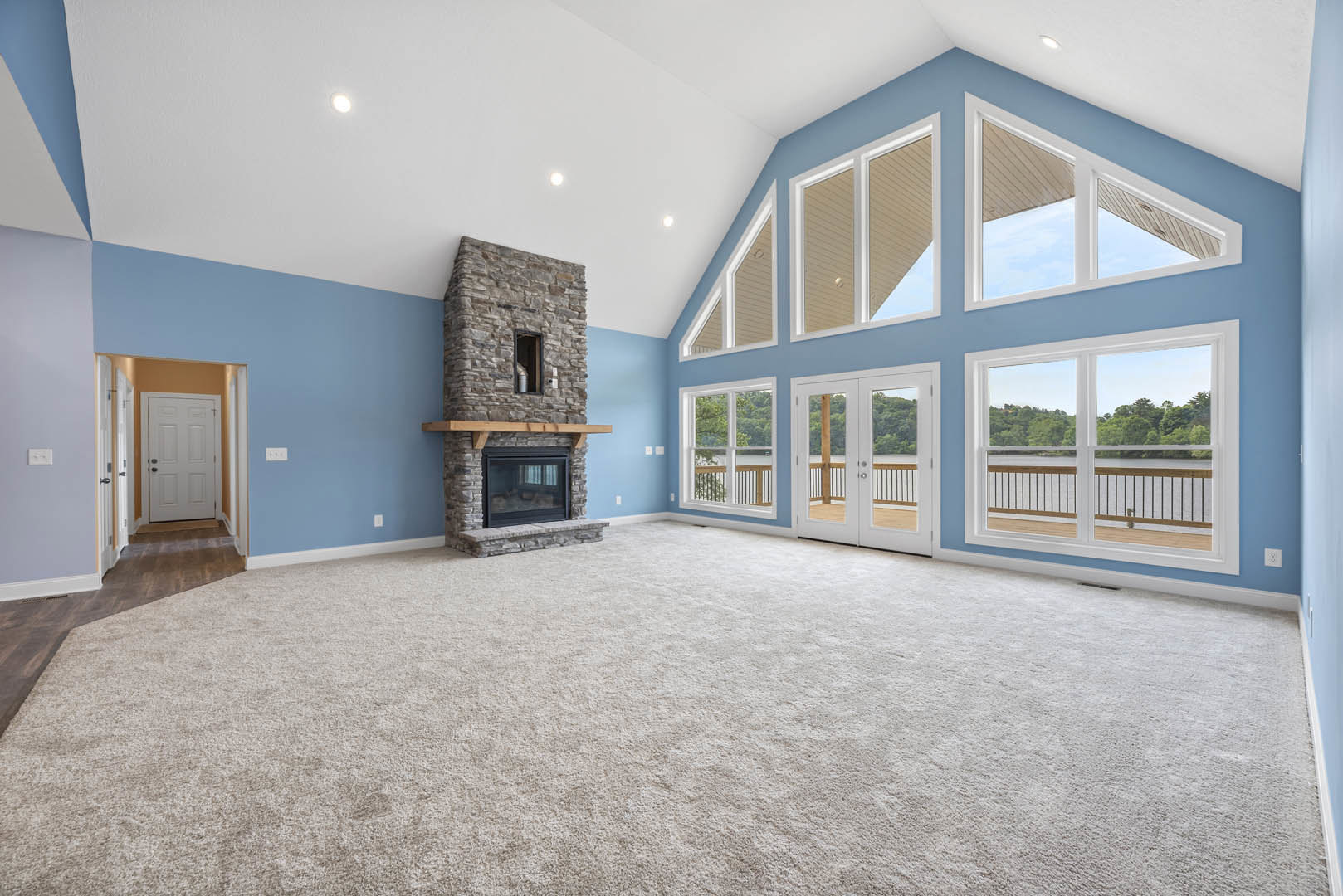 Spacious carpeted living room featuring a stone fireplace, large glass windows overlooking a lake and trees, and a white door with brown trim.
