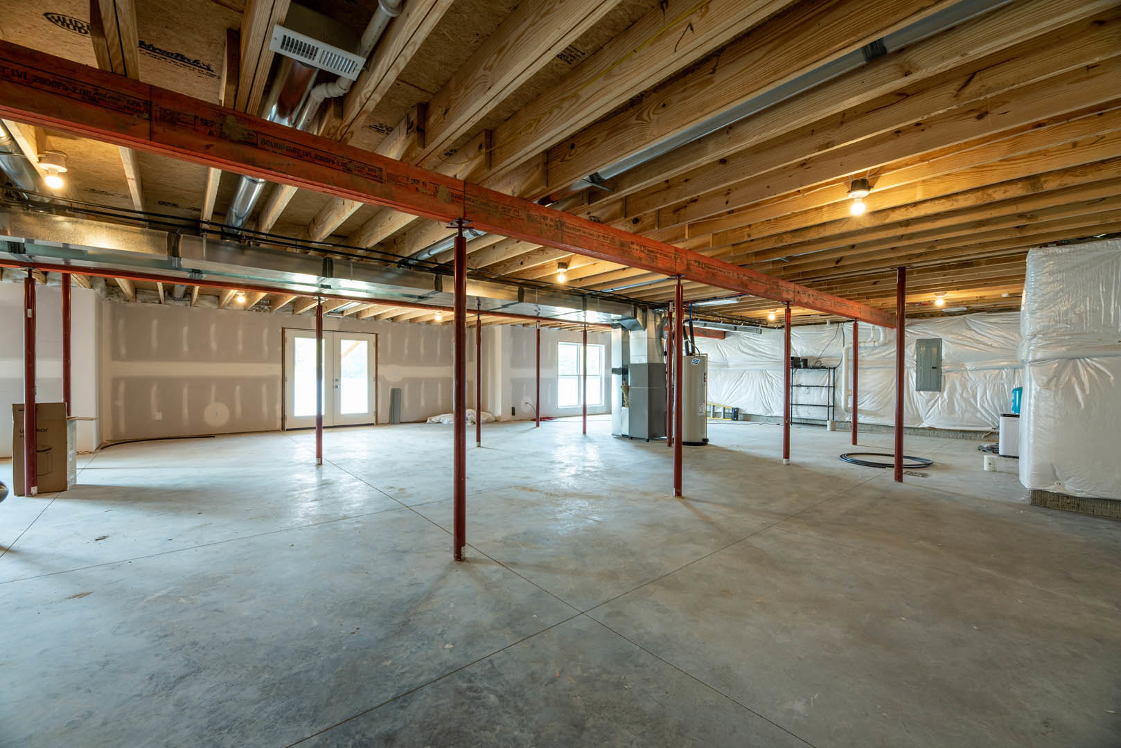 Exposed wooden ceiling beam above concrete floor in unfinished basement room with double doors and white glass door, red support poles, and light streaming through doorway