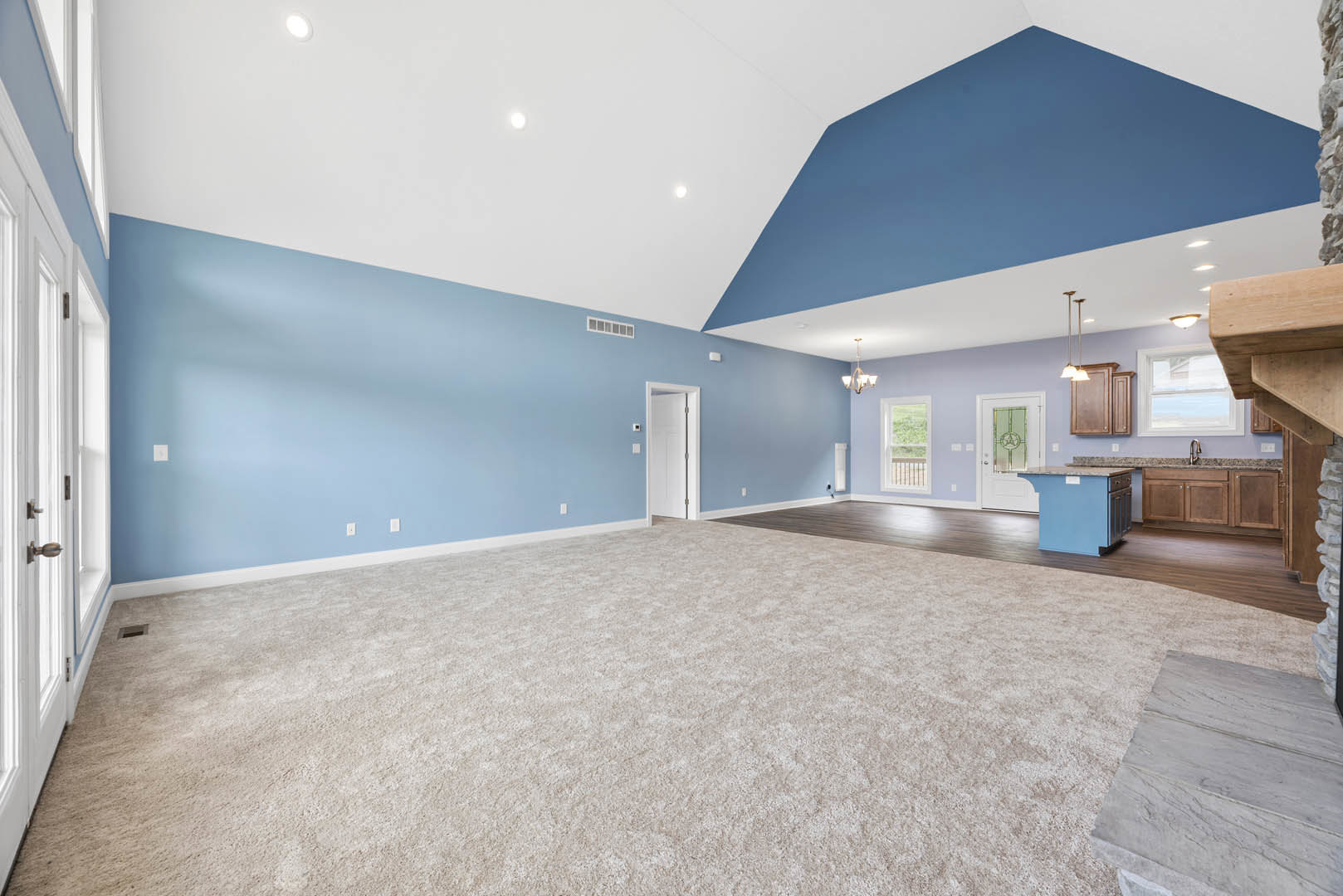 Open-concept kitchen and living area with high ceilings, carpeted floor, white and blue doors, large window overlooking grassy yard, close-up of stone countertop