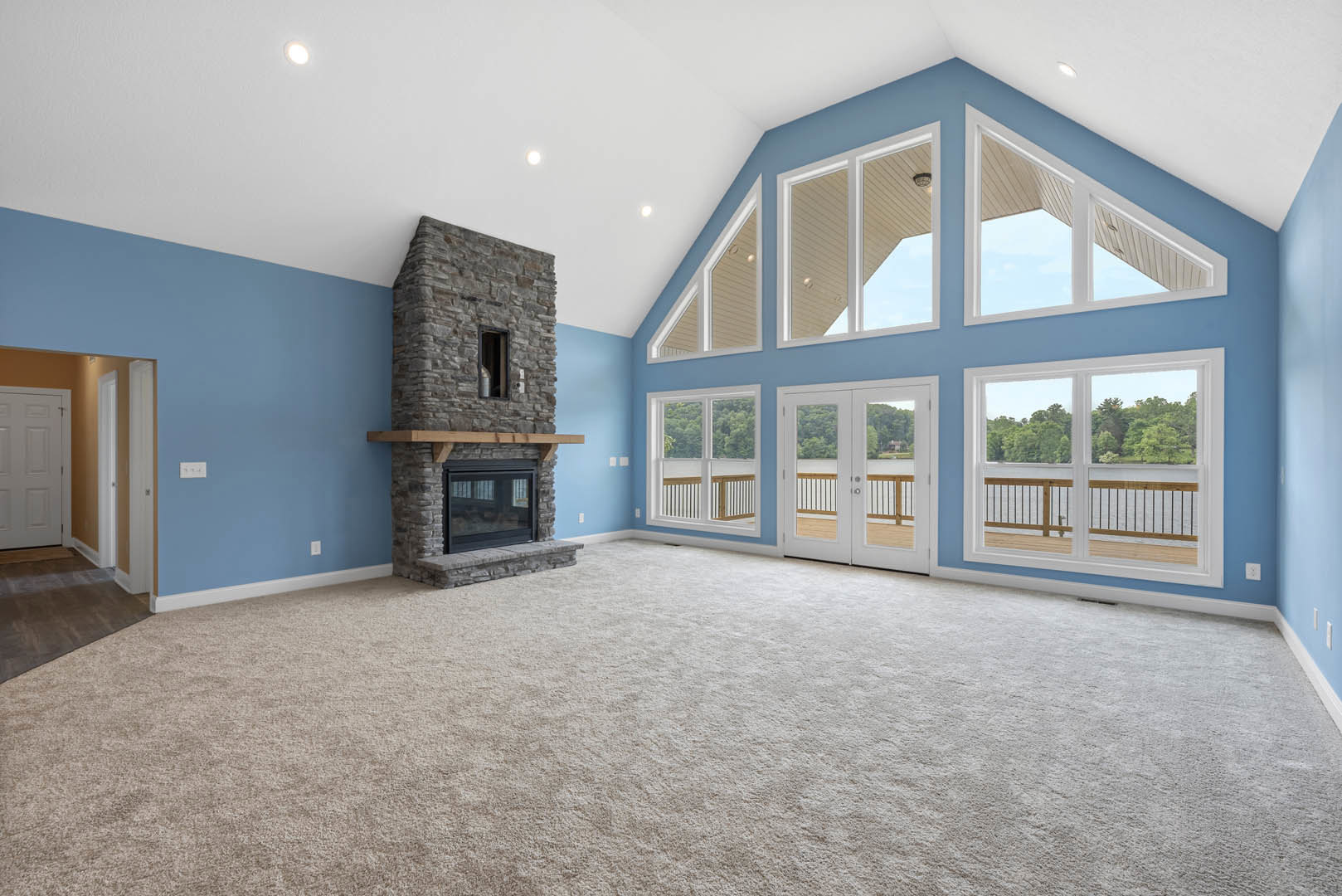 Stone fireplace with wood mantel, large glass windows, carpeted floor, white double doors with square glass panes, neutral walls and trim molding