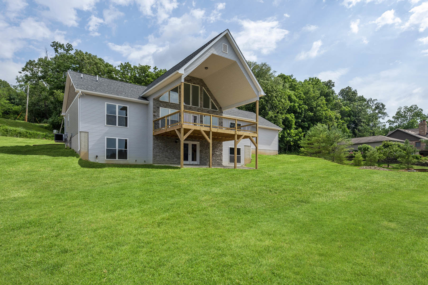 Two-story house with covered front porch, white siding, large windows, and manicured green lawn bordered by shrubs and trees under a partly cloudy sky