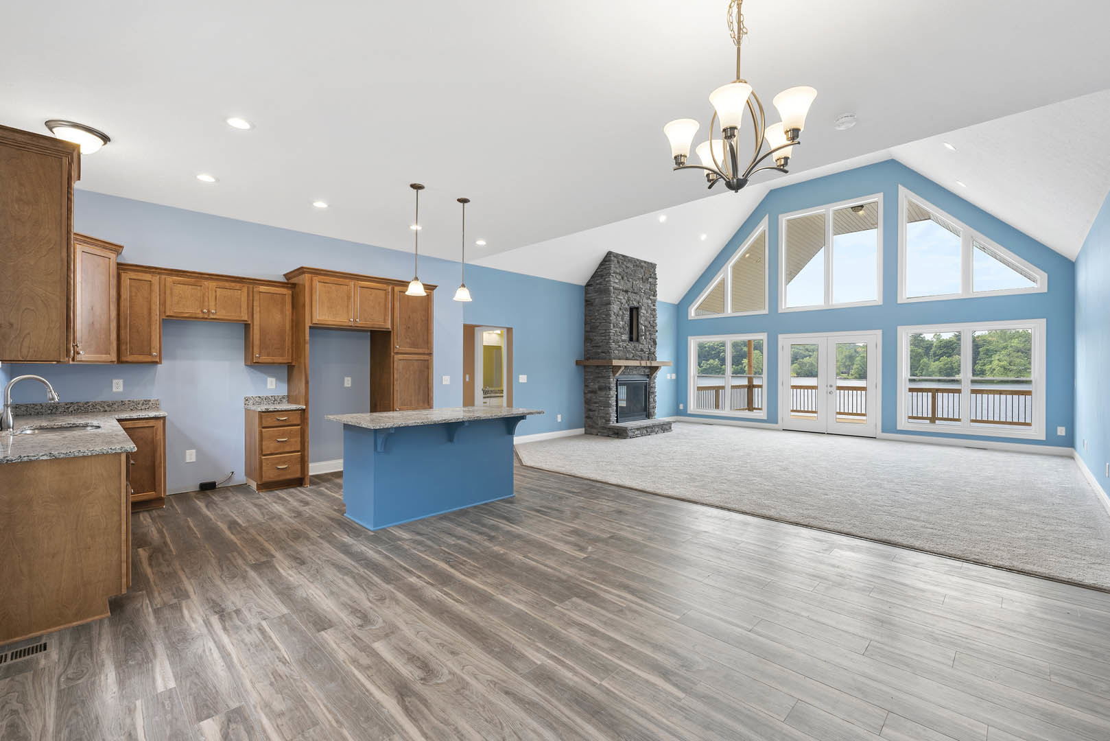 Spacious living room featuring a stone fireplace, tall ceiling, hardwood floors, blue kitchen counter with marble top, stone-framed window, and wooden dresser with marble surface