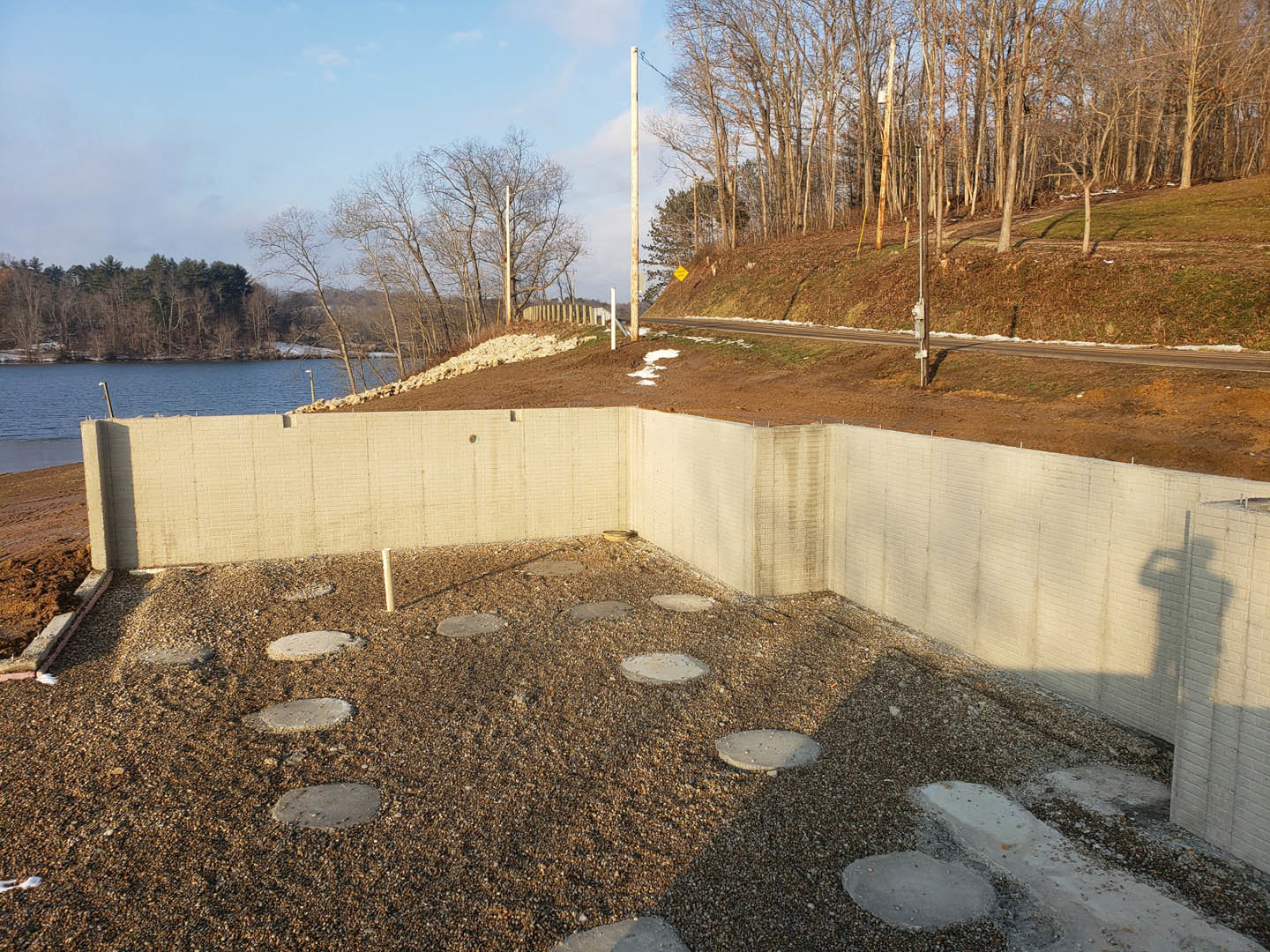 Stone retaining wall bordering a concrete patio, river and wooded hillside visible in the background, cloudy winter sky overhead.