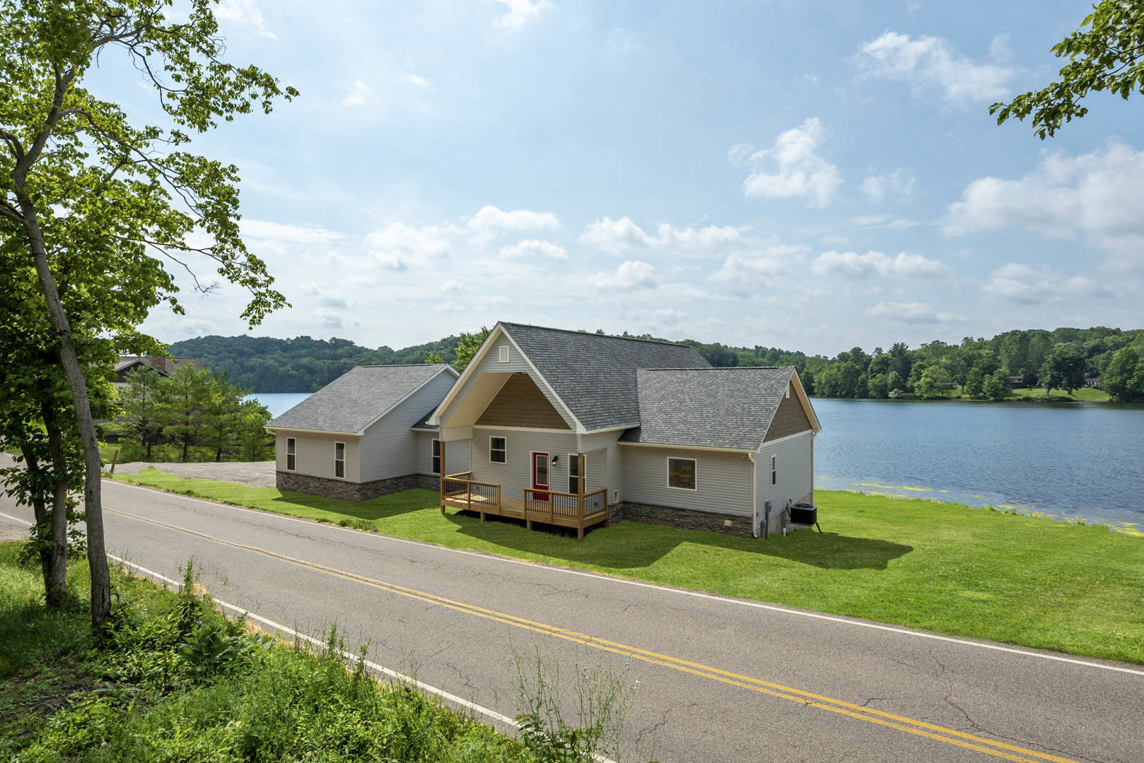 Two-story cottage with gray siding, red front door, and side deck with metal railing, situated beside a lake and surrounded by grass, trees, and a paved road.