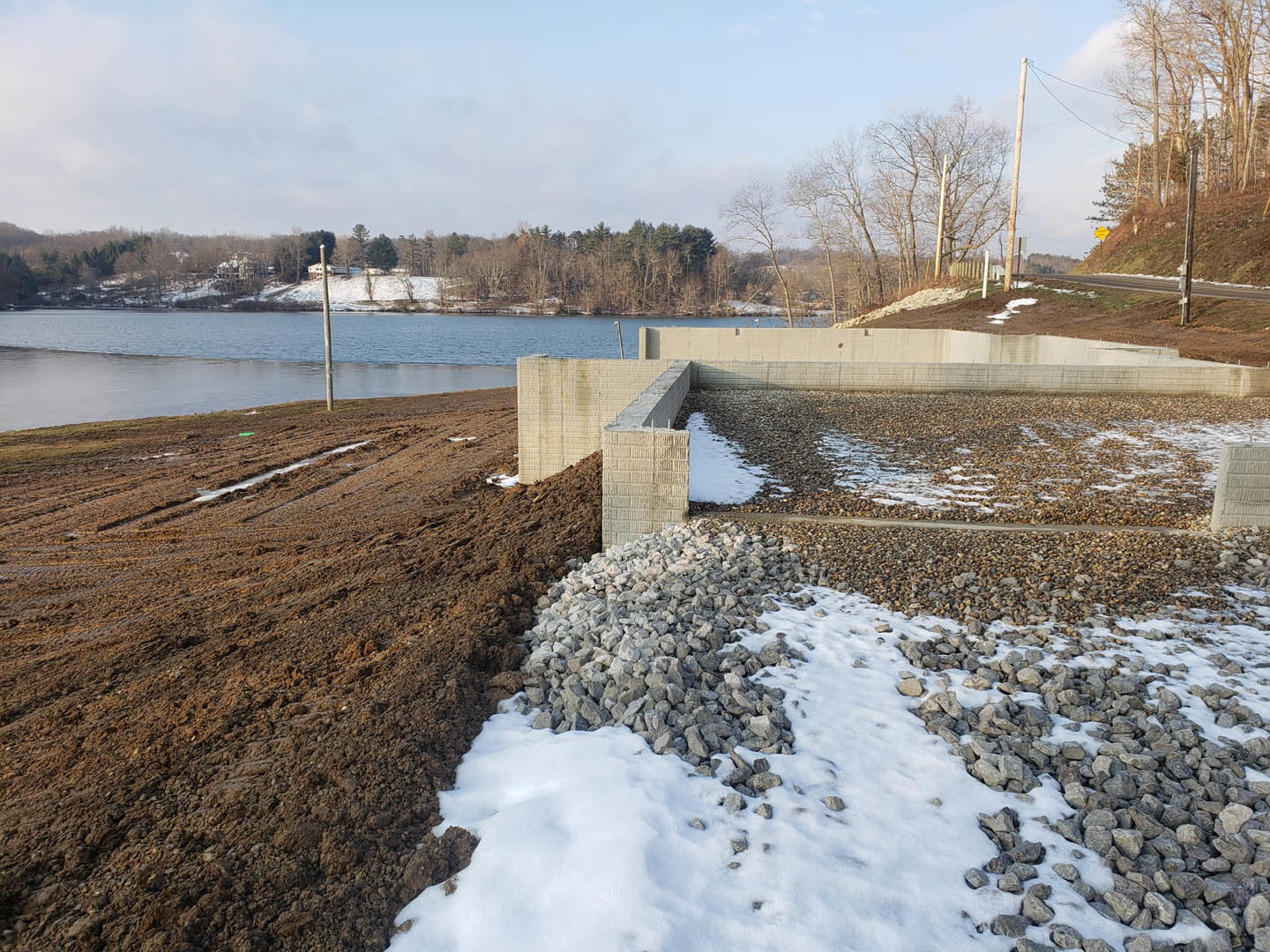 Snow-covered ground and rocks beside a lake, with a muddy field, scattered trees, and blue sky with clouds in the background