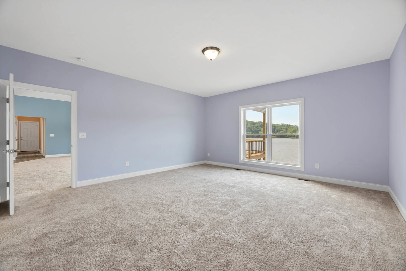 Carpeted room with large window overlooking a lake, white door in hallway, ceiling light fixture, and wooden railing in foreground