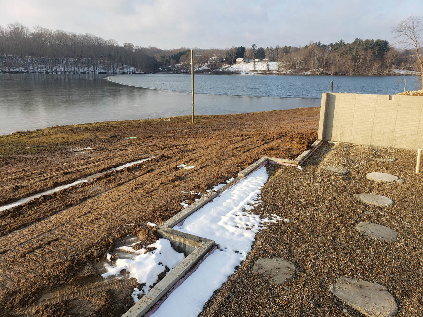 Snow-covered ground beside a lake, bare trees along the shoreline, blue sky with scattered clouds, white brick wall visible near the water, grassy area with a pole in the