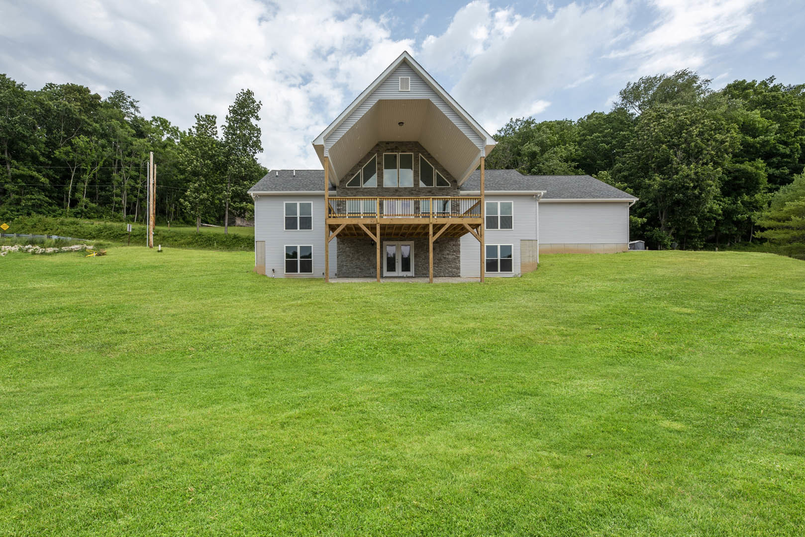 Two-story house with white double doors and glass panes, wood deck porch, upper balcony, large green lawn, white-framed windows, gray roof, surrounded by trees and plants under a