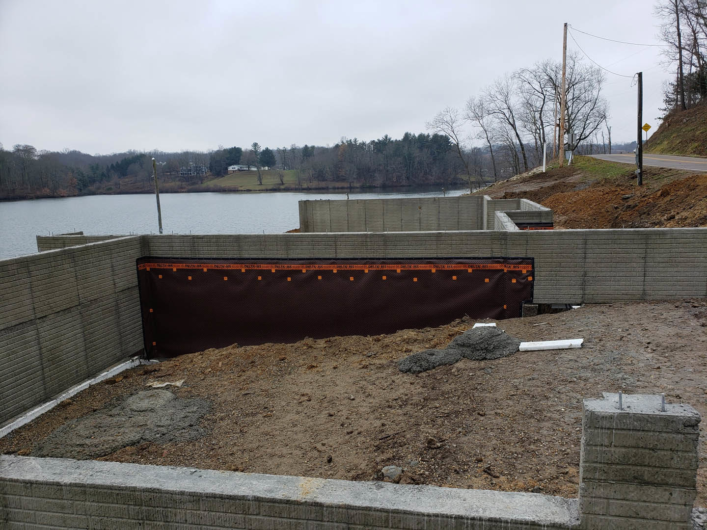 Dirt and rocks surrounded by fencing at a lakeside construction site, black retaining wall marked with orange tape, concrete block with metal bar, distant trees under a cloudy sky
