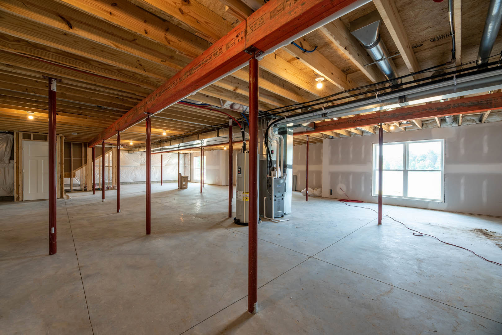 Exposed pipes along ceiling and walls, large window with tree view, red support pole beside wooden beam, unfinished floor and walls in spacious room