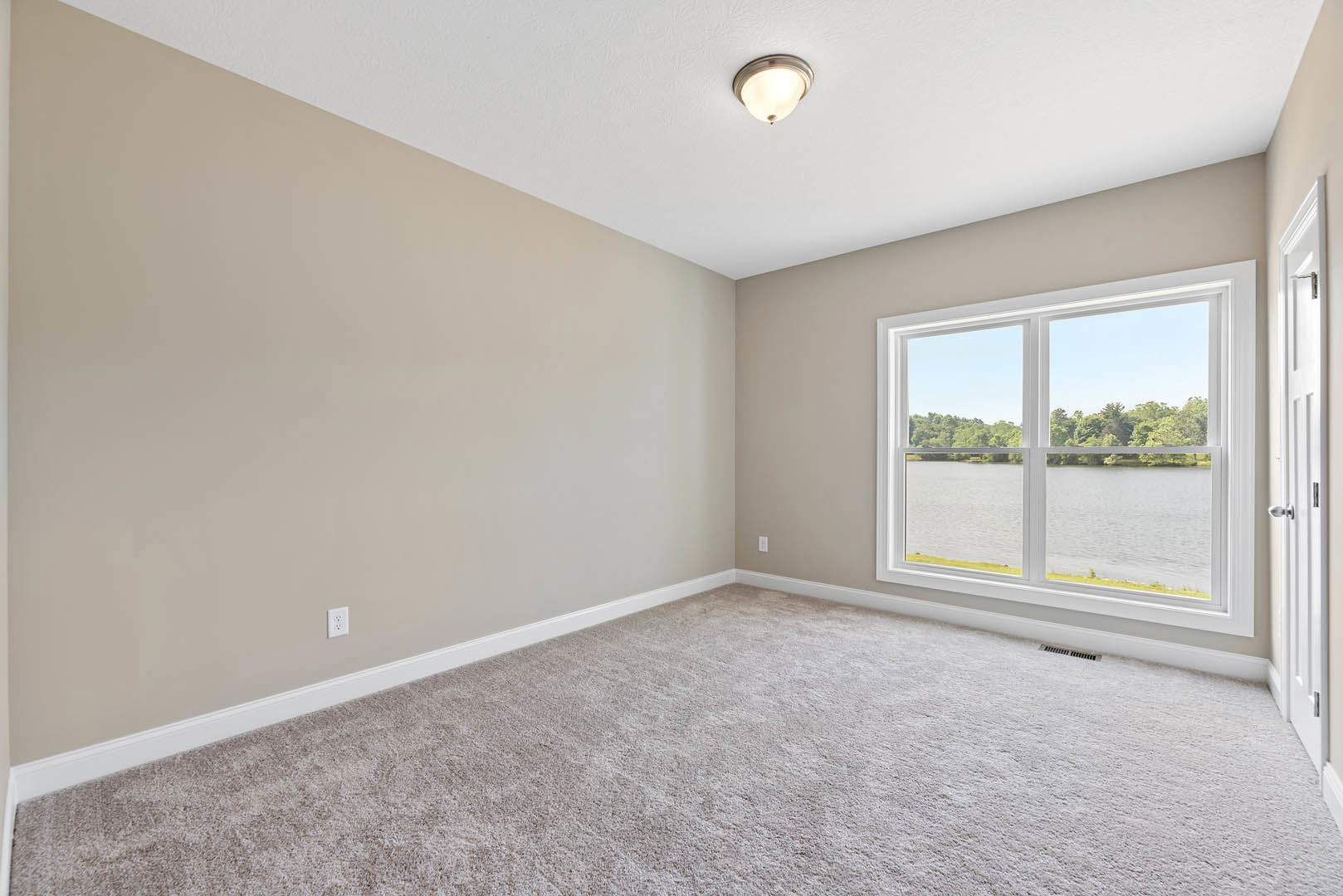 Carpeted room with white walls, ceiling light fixture, large window overlooking trees and lake, close-up of wooden door, decorative molding along ceiling and floor.