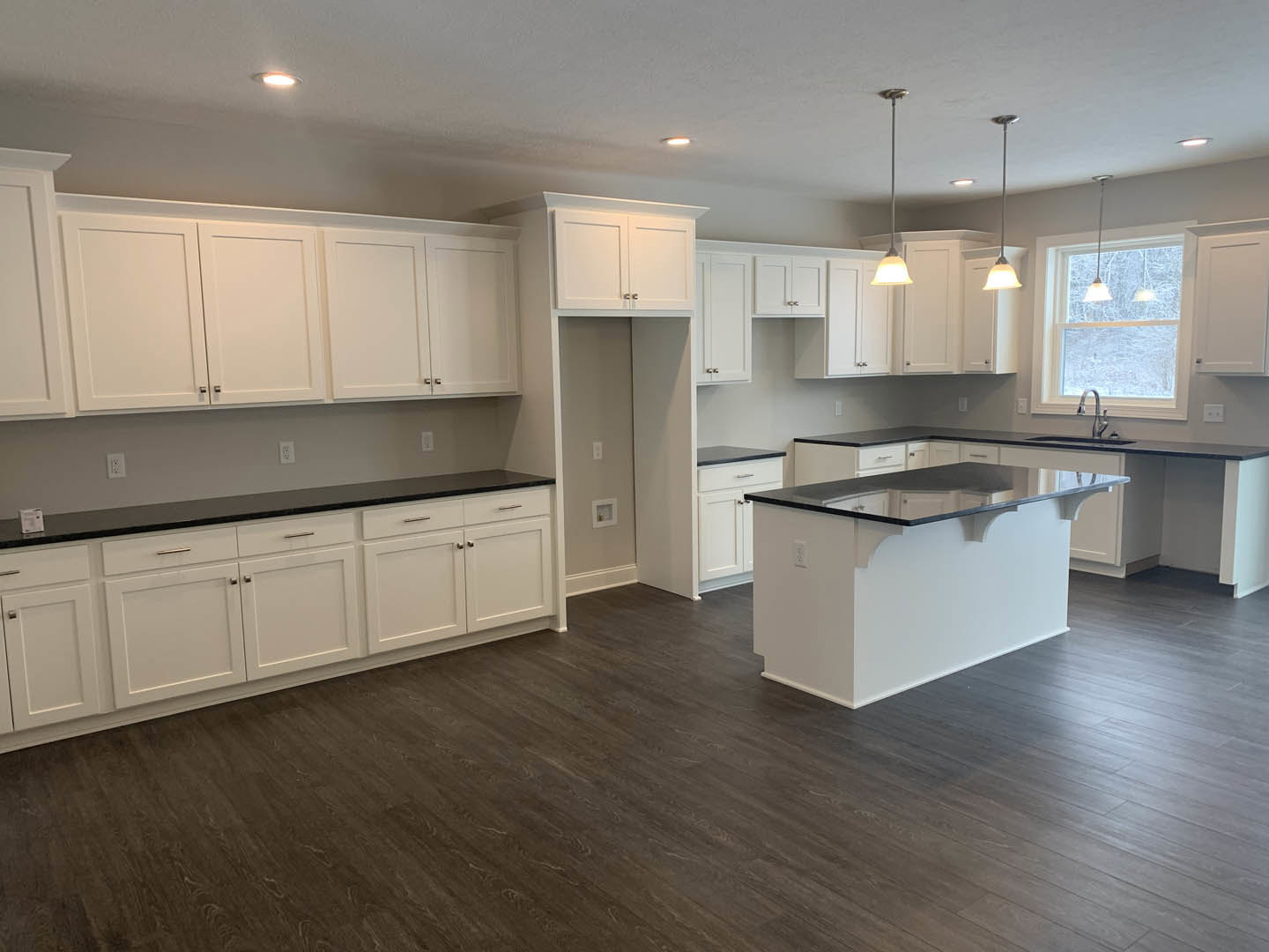 White kitchen cabinets with black countertops, dark wood flooring, kitchen island with matching black surface, stainless steel sink, white door, large window with a woman seated in