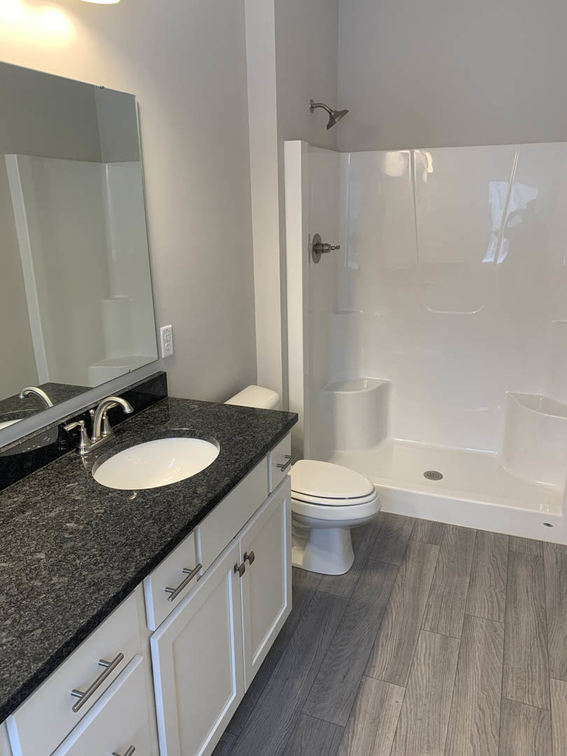Modern bathroom featuring a granite countertop with undermount sink, wall-mounted mirror, white tiled shower enclosure with glass door, and adjacent toilet.
