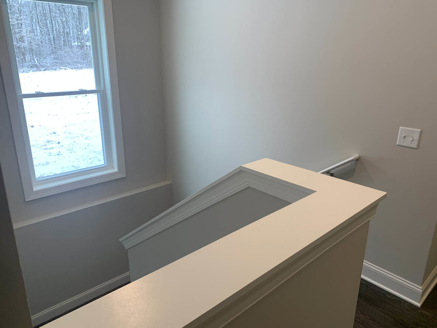 White staircase with wood treads beside a large window overlooking snowy forest, white walls and light switch visible