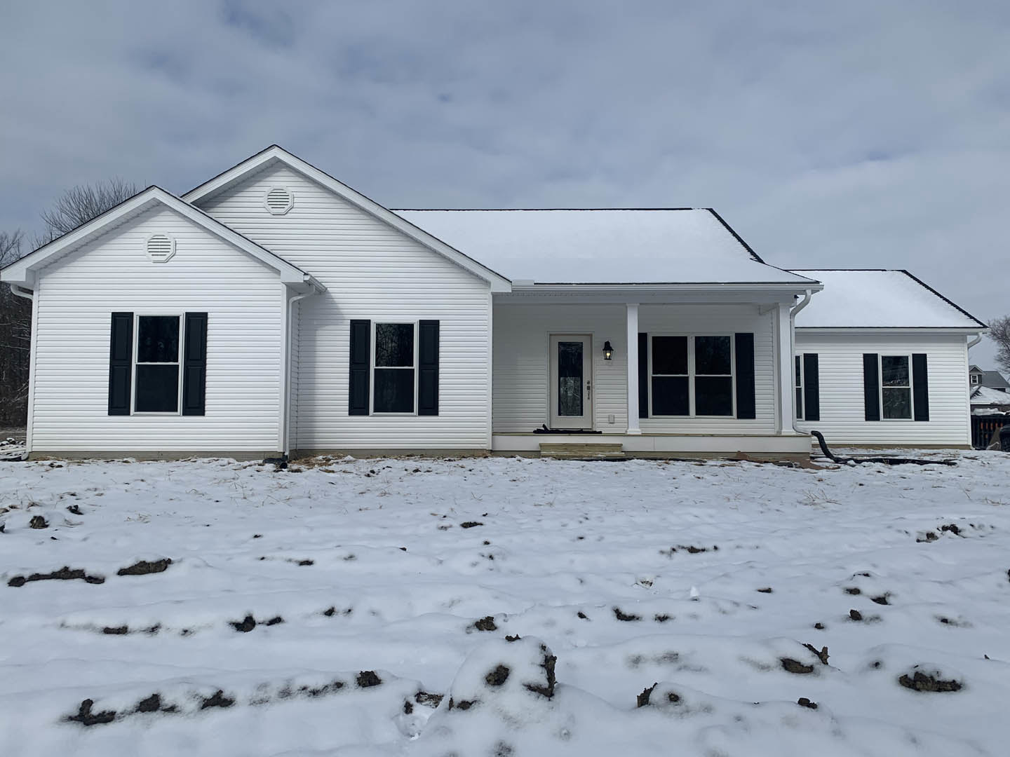 White house with black shuttered windows, glass panel front door, snow covering roof and ground, winter landscape under cloudy sky