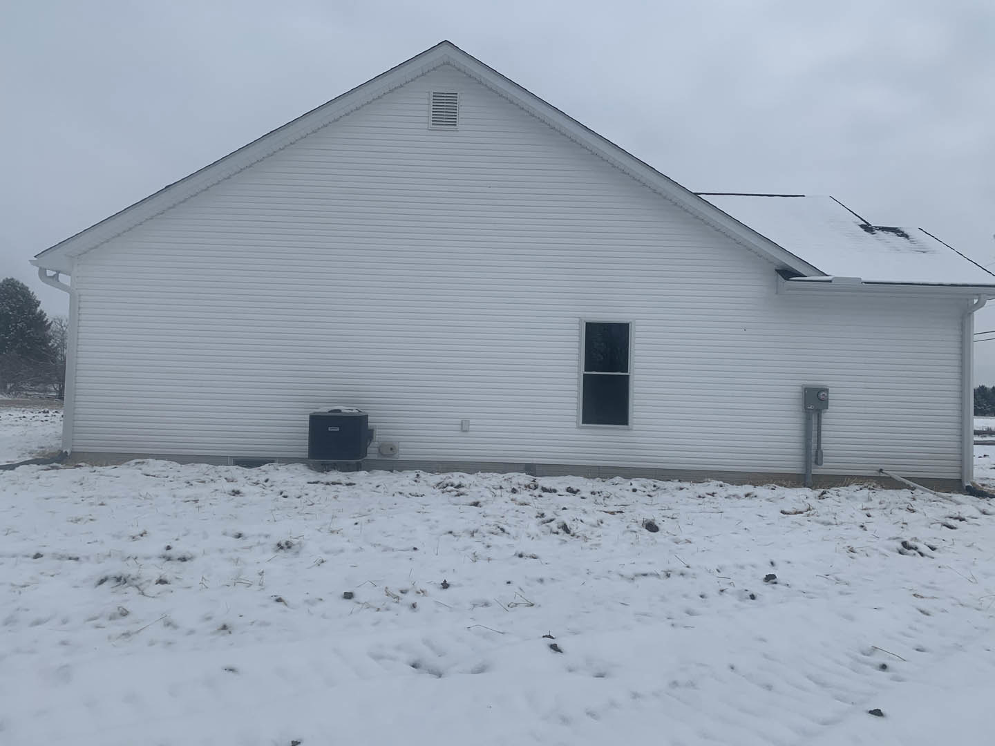White house with horizontal siding, rectangular window with white trim, snow covering ground with patches of grass and dirt, winter sky in background