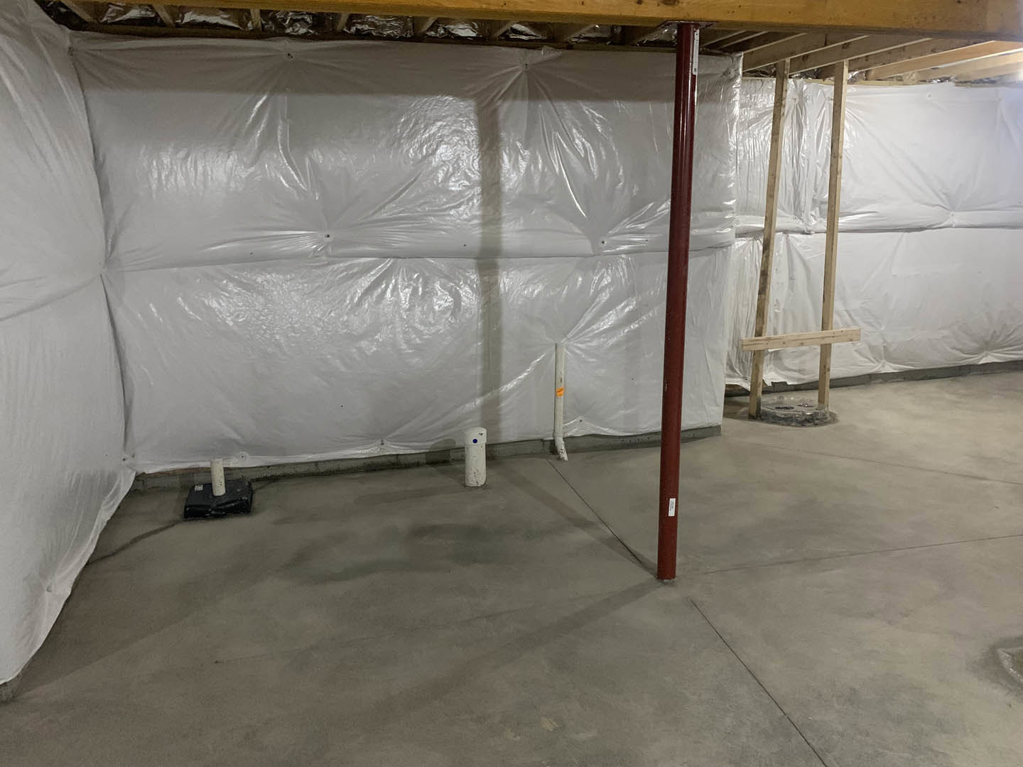 White plastic sheeting covers a wall above a concrete floor, with a black box mounted on a white pole, red poles nearby, a close-up of a pipe, a wooden ladder, and a white sheet of
