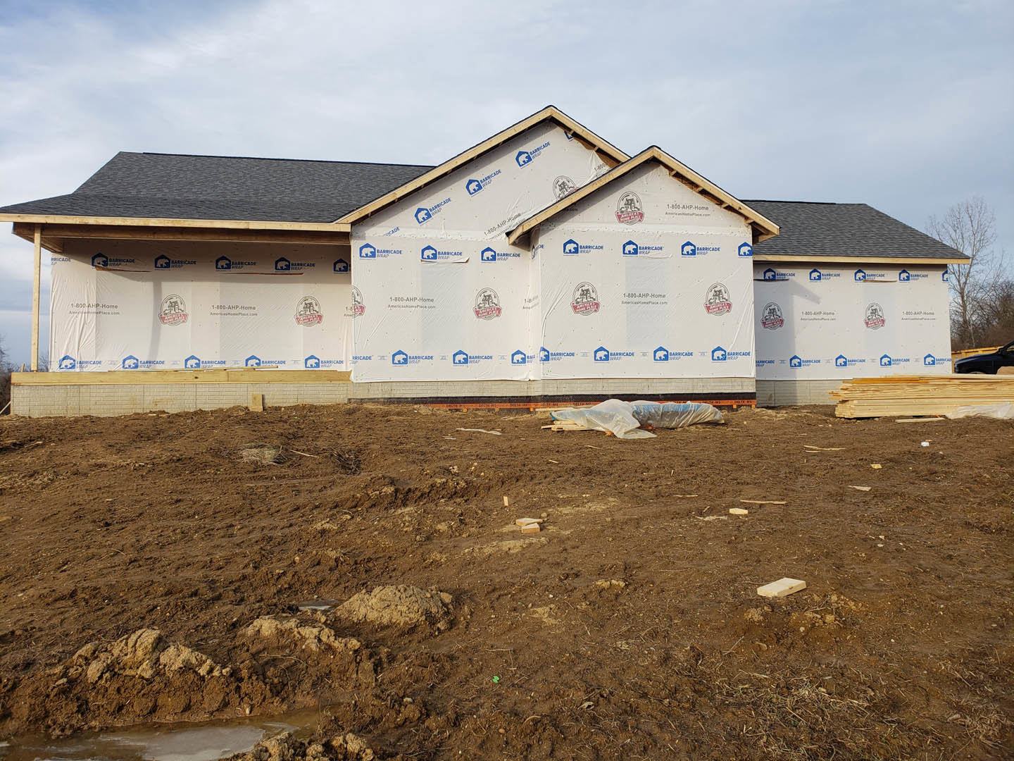 Two-story house under construction with white protective sheets covering the roof, muddy ground in front, pile of dirt, and scattered construction materials.