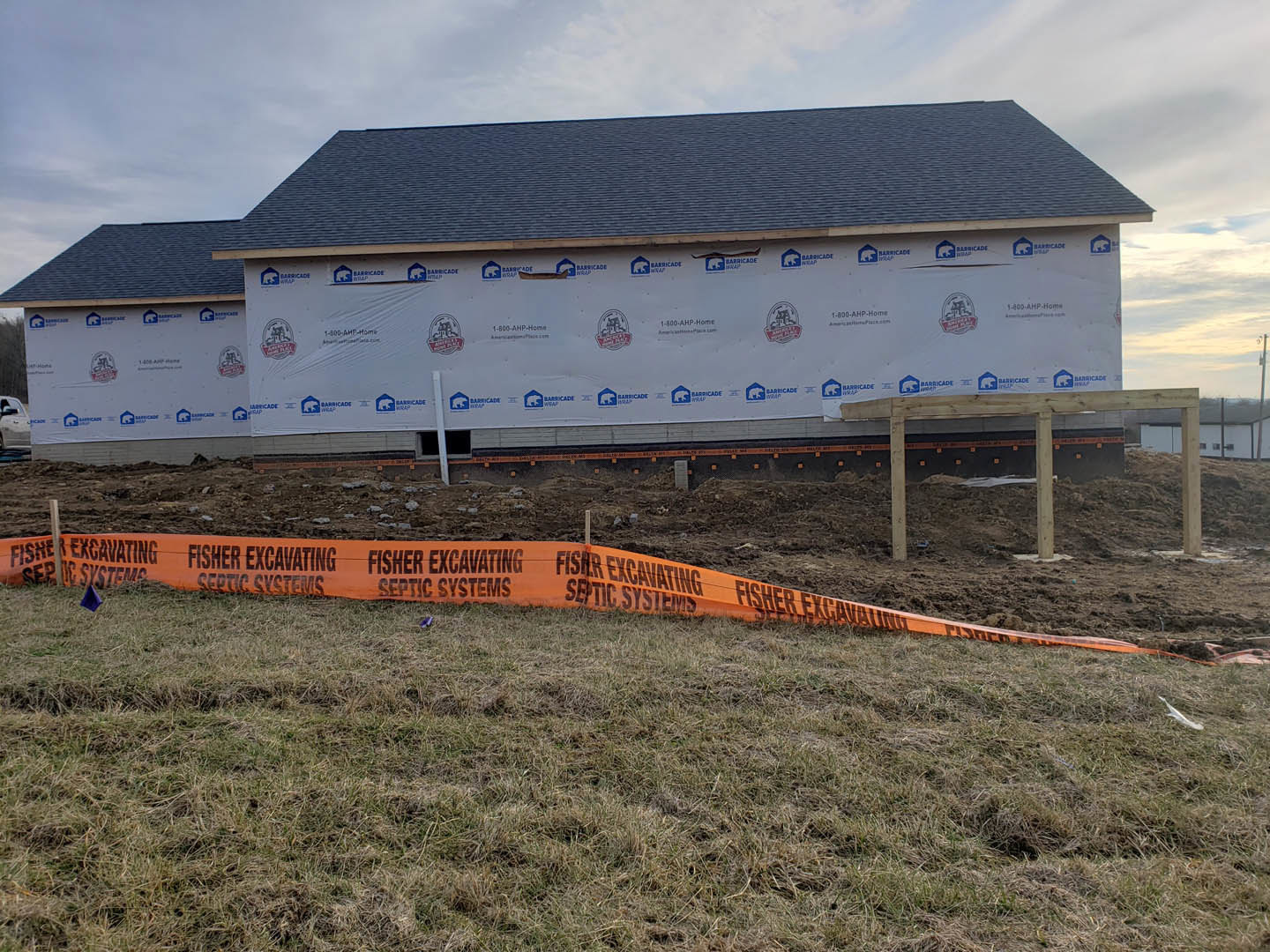 Framed custom home under construction on grassy lot, orange safety tape marking perimeter, cloudy sky overhead