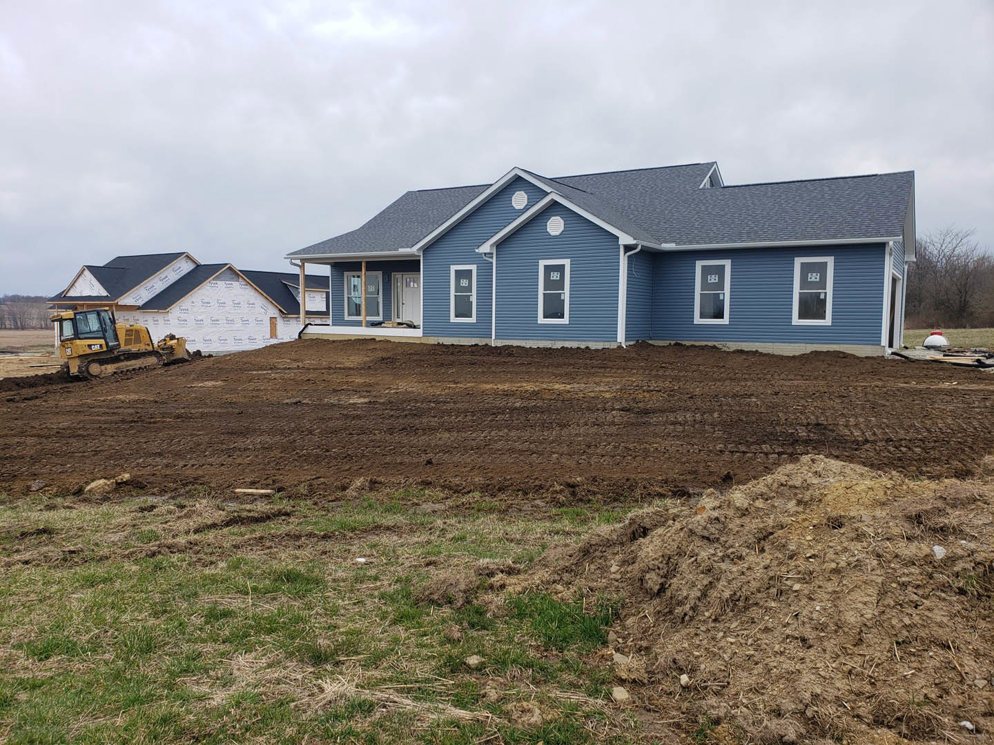 Blue house with white trim, large windows, dirt field and pile in front, yellow bulldozer parked at construction site, cloudy sky overhead