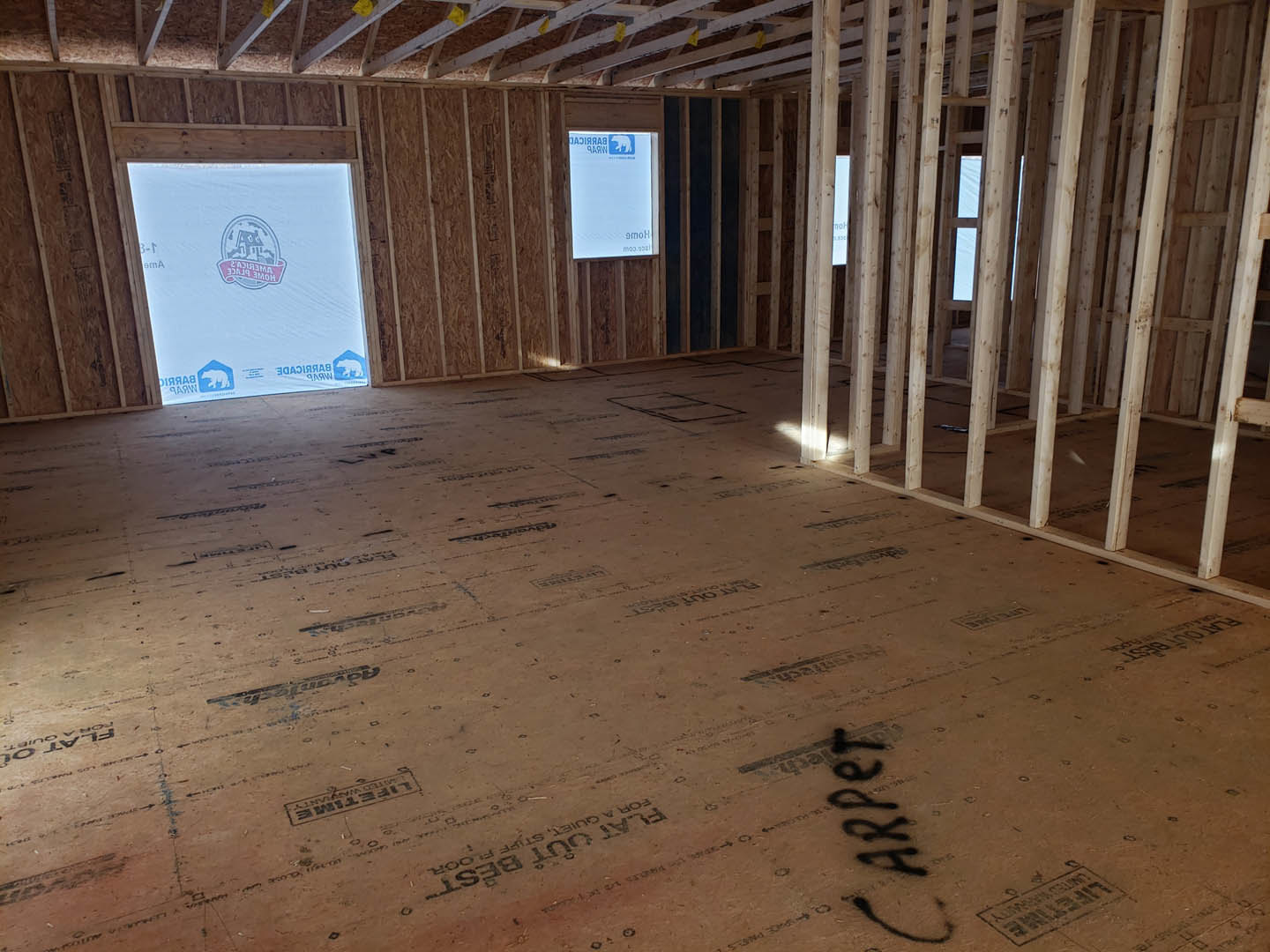 Exposed wood ceiling beams, unfinished floor, large window with a white and blue sign, construction materials visible in a residential interior.