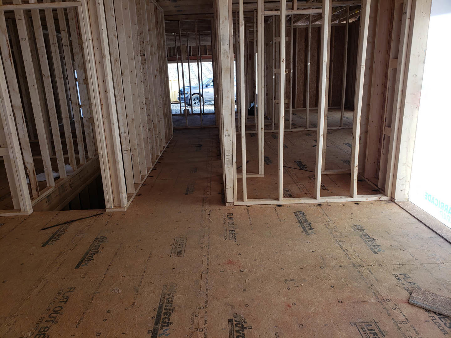 Room under construction with exposed wood framing, plywood floor marked with black text, white wall featuring a central light fixture, wooden door with glass window, and parked car