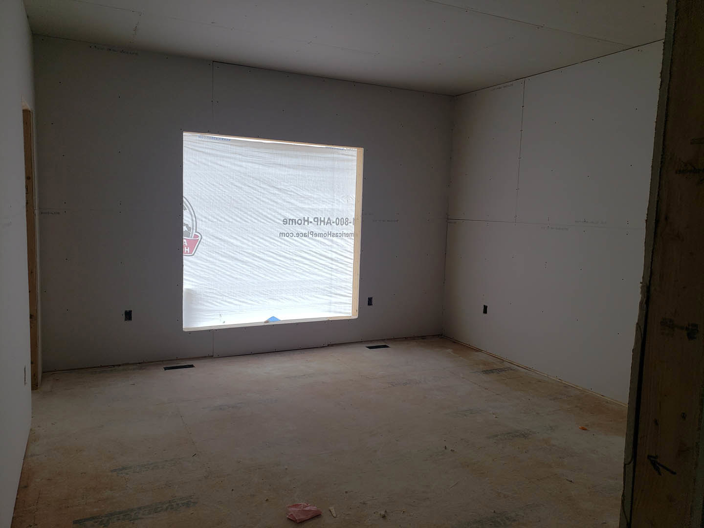 Bright room with large window, white plastered walls, folded red cloth on a surface, unfinished floor, and exposed ceiling.