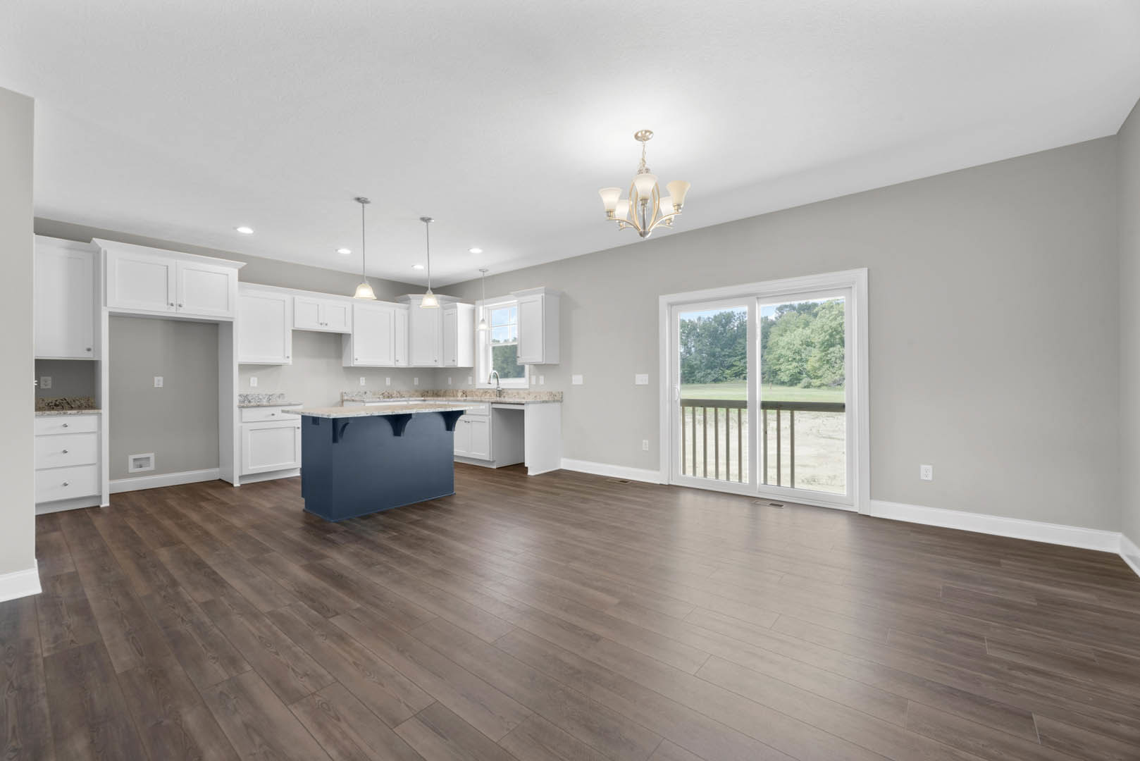 Spacious kitchen and living area featuring hardwood floors, blue kitchen island with marble countertop, sliding glass door overlooking a field of trees, white-framed window, and