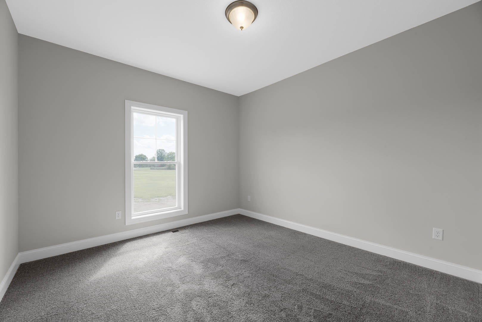 Bright room with a large window overlooking a grassy field, grey carpet flooring, white walls, ceiling light fixture, and simple molding trim