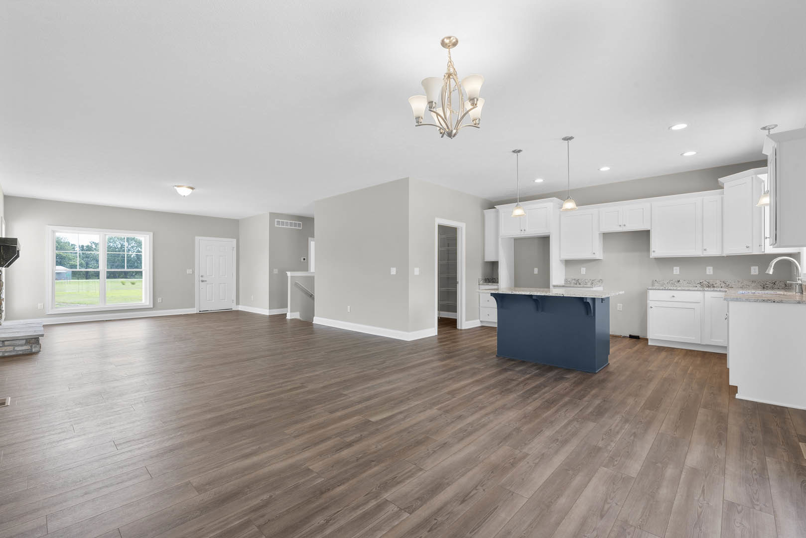 Spacious open floor plan featuring a kitchen island with white countertop, hardwood flooring throughout, blue accent wall with built-in white shelves, large window overlooking