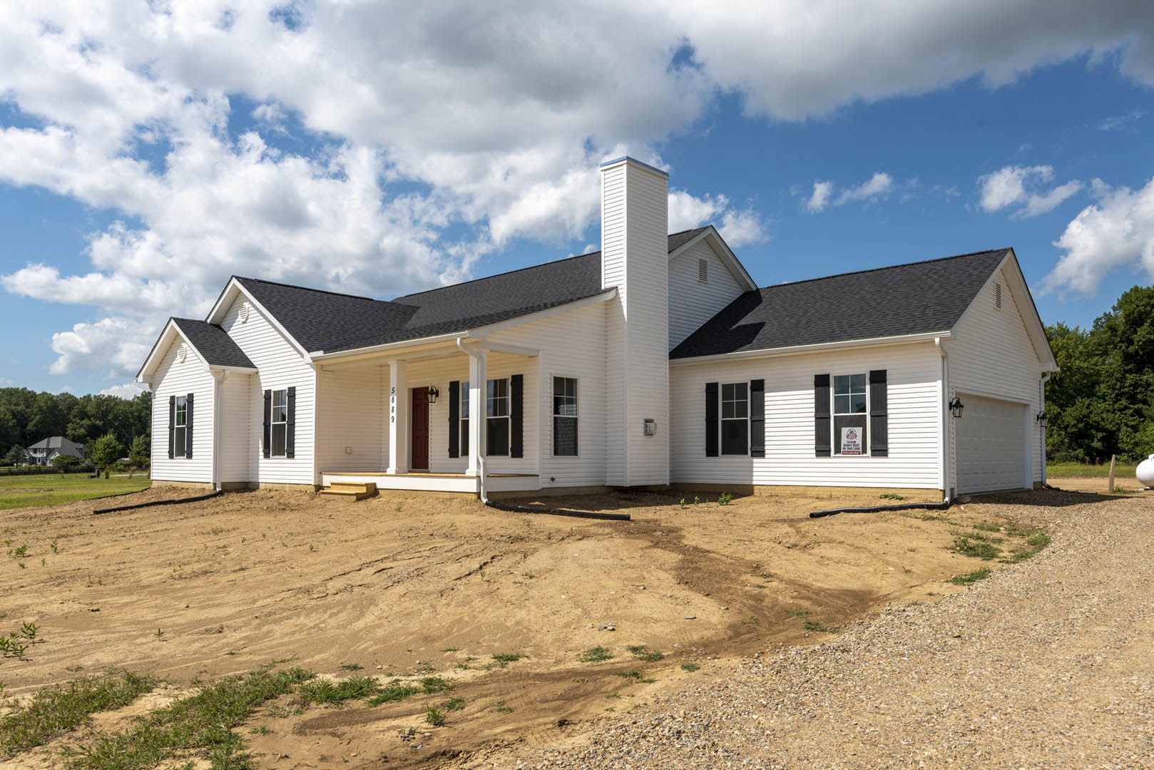 White house with black roof and chimney, dirt hill in front, large front window displaying a sign, surrounded by sparse trees under cloudy sky