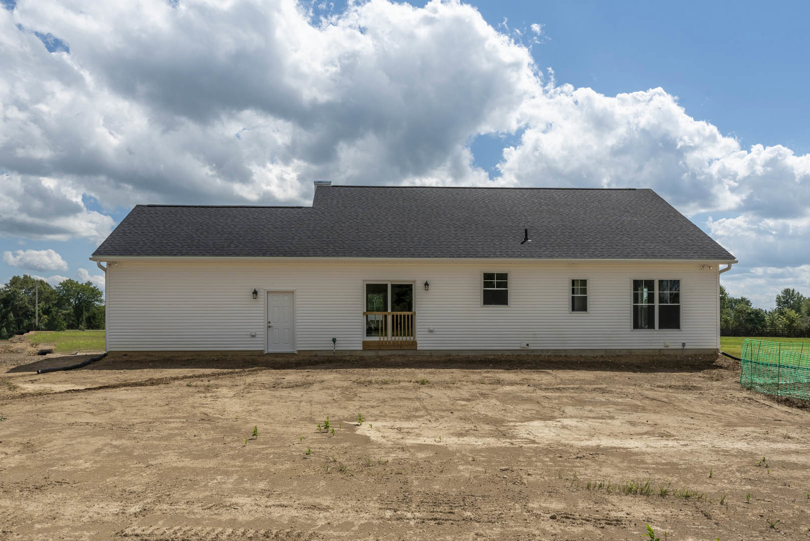 White house with covered porch, white door with silver handle, green mesh fence bordering a dirt yard with small plants, cloudy sky above pitched roof.