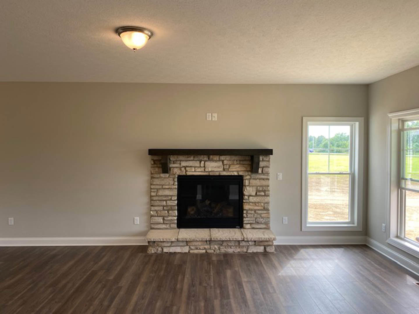 Modern living room with black glass fireplace, stone accent wall, wide wood plank flooring, large window overlooking grassy yard and trees, and recessed ceiling light fixture