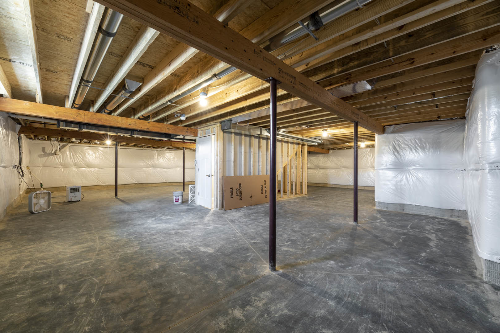 White wall with exposed wooden ceiling beams, visible pipes, concrete floor, large cardboard box, small metal fan, black wire, and plastic-wrapped insulation along the wall.