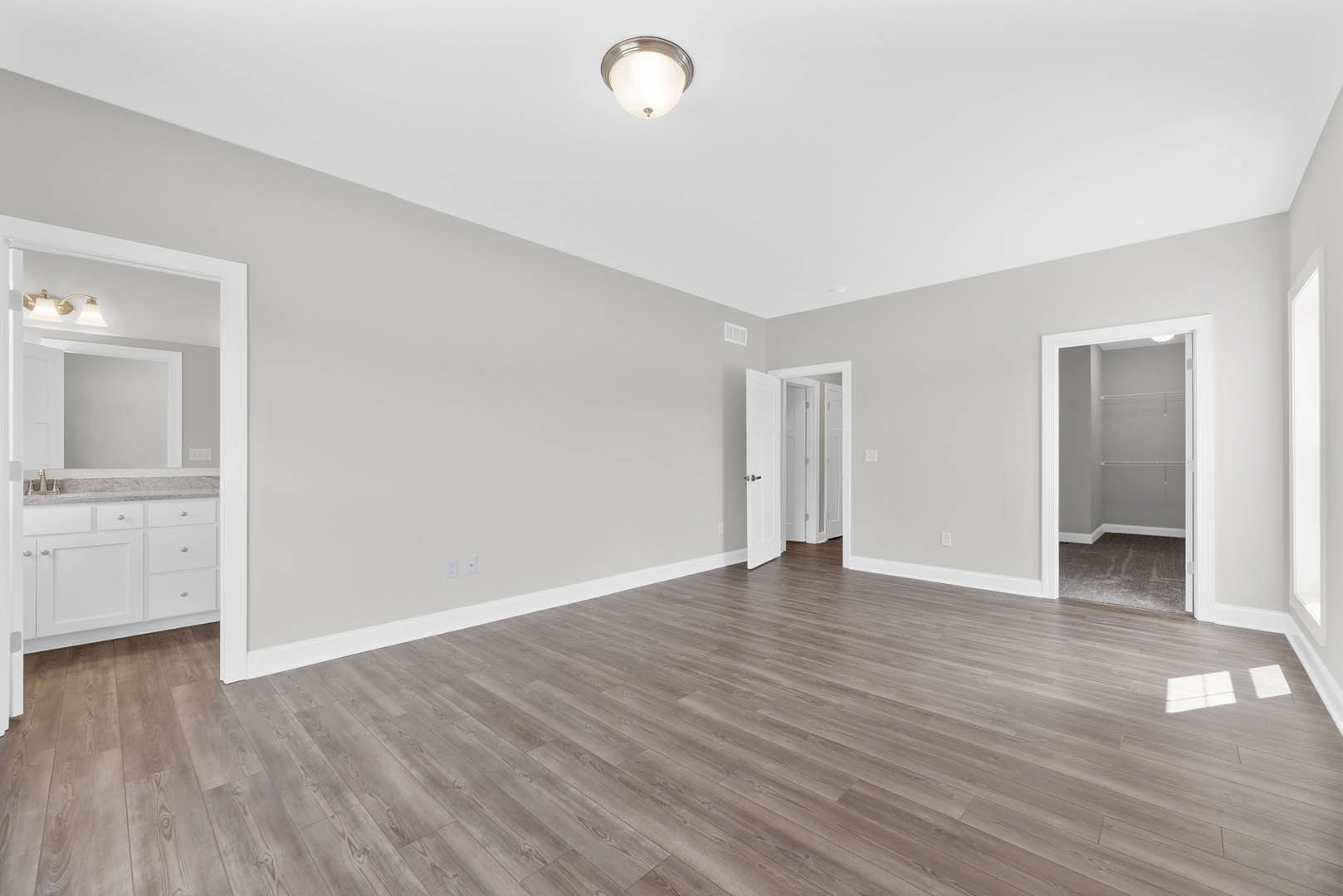 Open room with hardwood flooring, white walls, white baseboard trim, and a glimpse of a bathroom featuring white cabinetry and a wall mirror