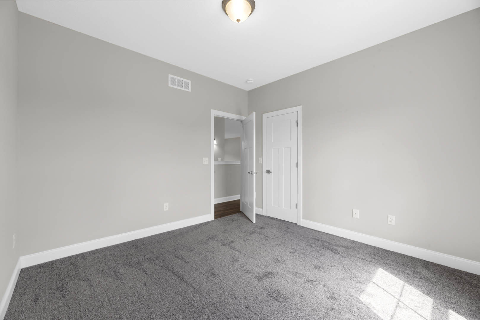 White paneled door with silver knob open onto grey carpeted room, white walls with wall vent, ceiling molding, and modern wall light fixture