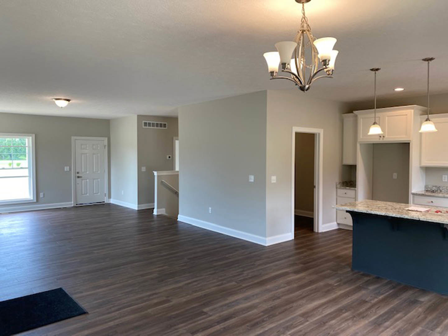 Open kitchen and dining area with marble-topped island, wood flooring, black mat, four-shade ceiling light fixture, white door with silver handle, plaster walls, and laminate