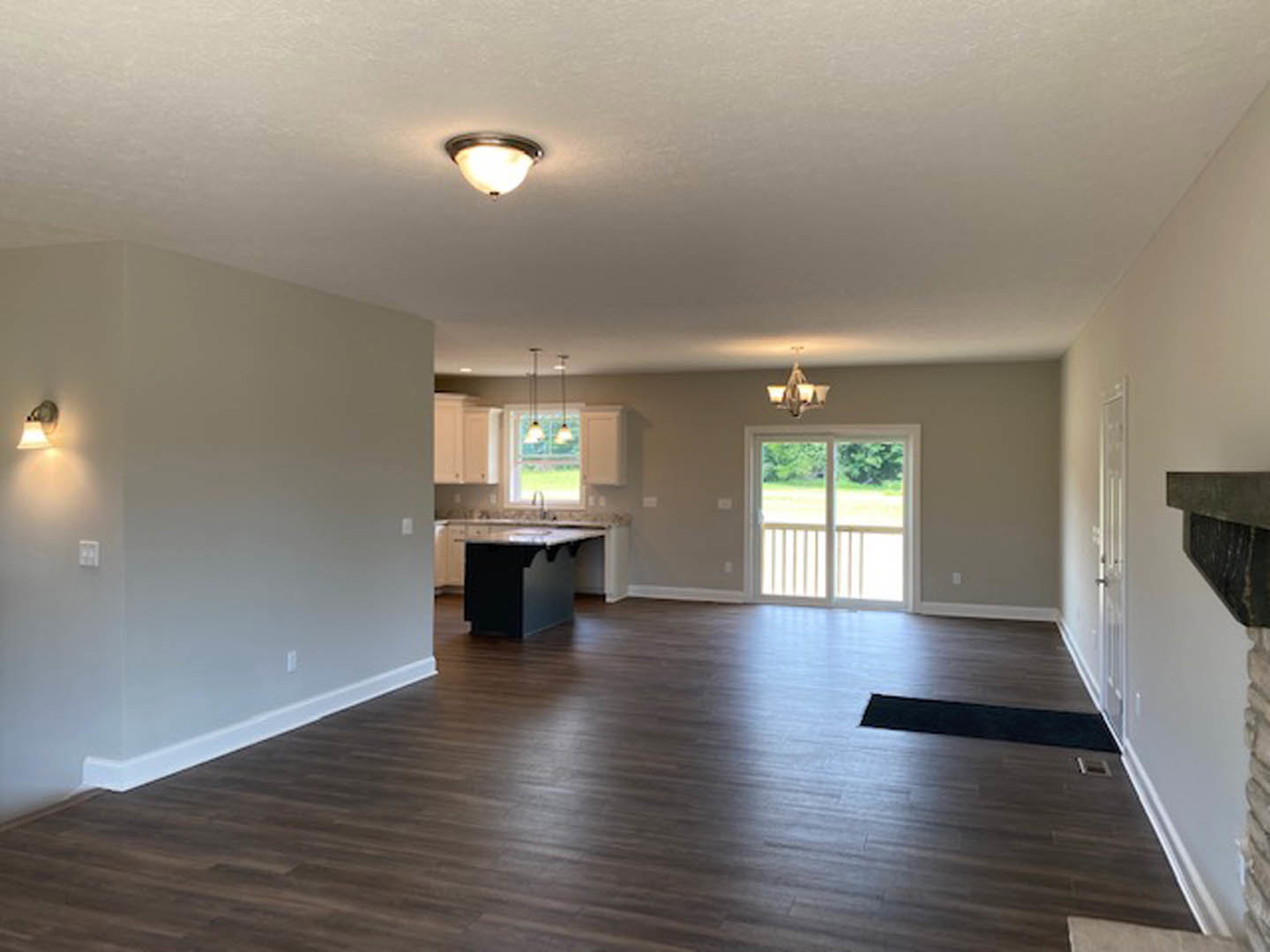Spacious open-plan room featuring wood flooring, a kitchen with a black island topped with marble, adjacent dining area, white sliding glass door, wall-mounted light fixture, and a