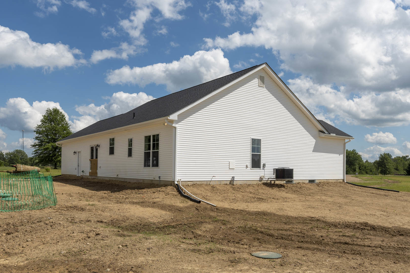 White house with black roof, green netting on dirt field, exposed pipe, blue sky with scattered clouds