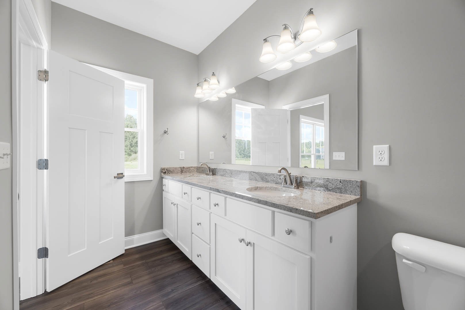 Bathroom with double vanity featuring undermount sinks, quartz countertop, large framed mirror, row of chrome light fixtures above, white cabinetry, wood-look tile flooring, and