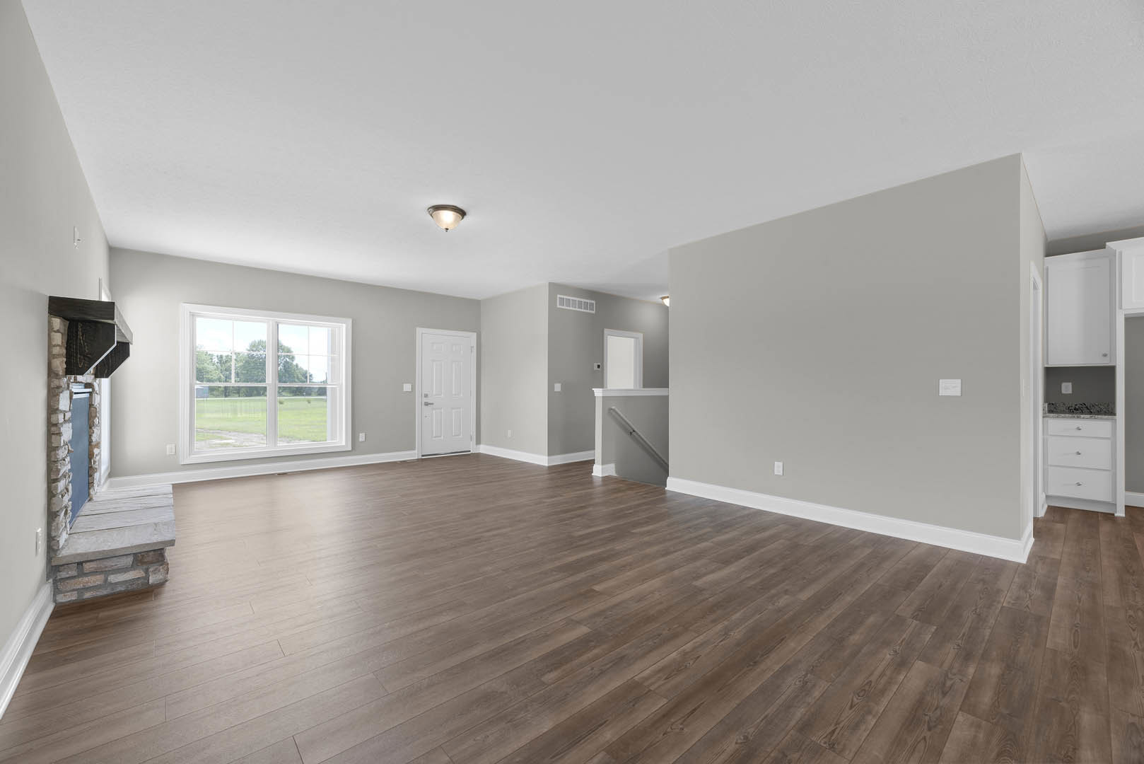 Wood flooring in a bright room with a large window overlooking a grassy field, white walls, ceiling light fixture, and a white door with silver handles.