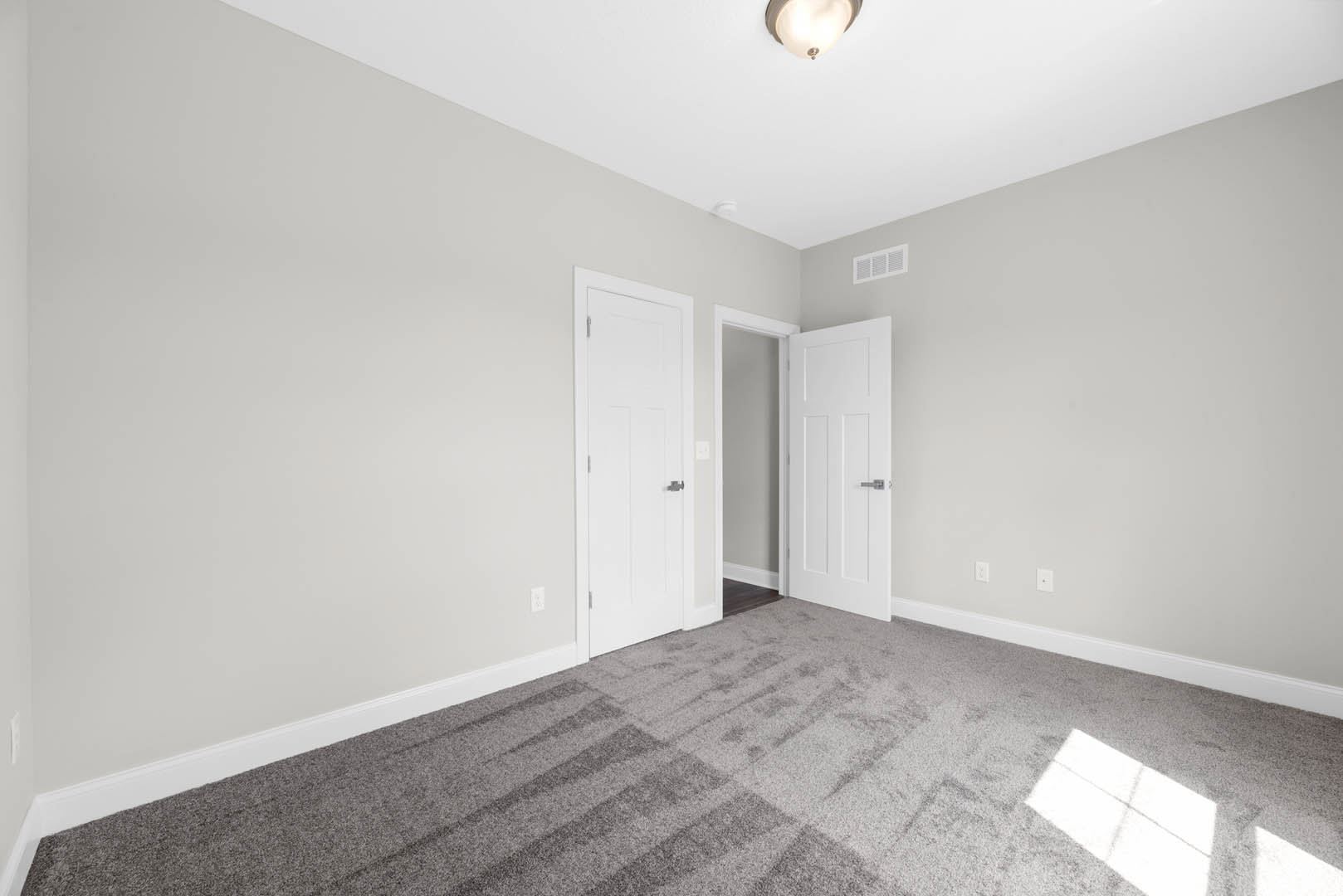 Carpeted room with two white doors, one featuring a black handle and the other a silver handle, white walls with base molding, ceiling light fixture, and wall vent
