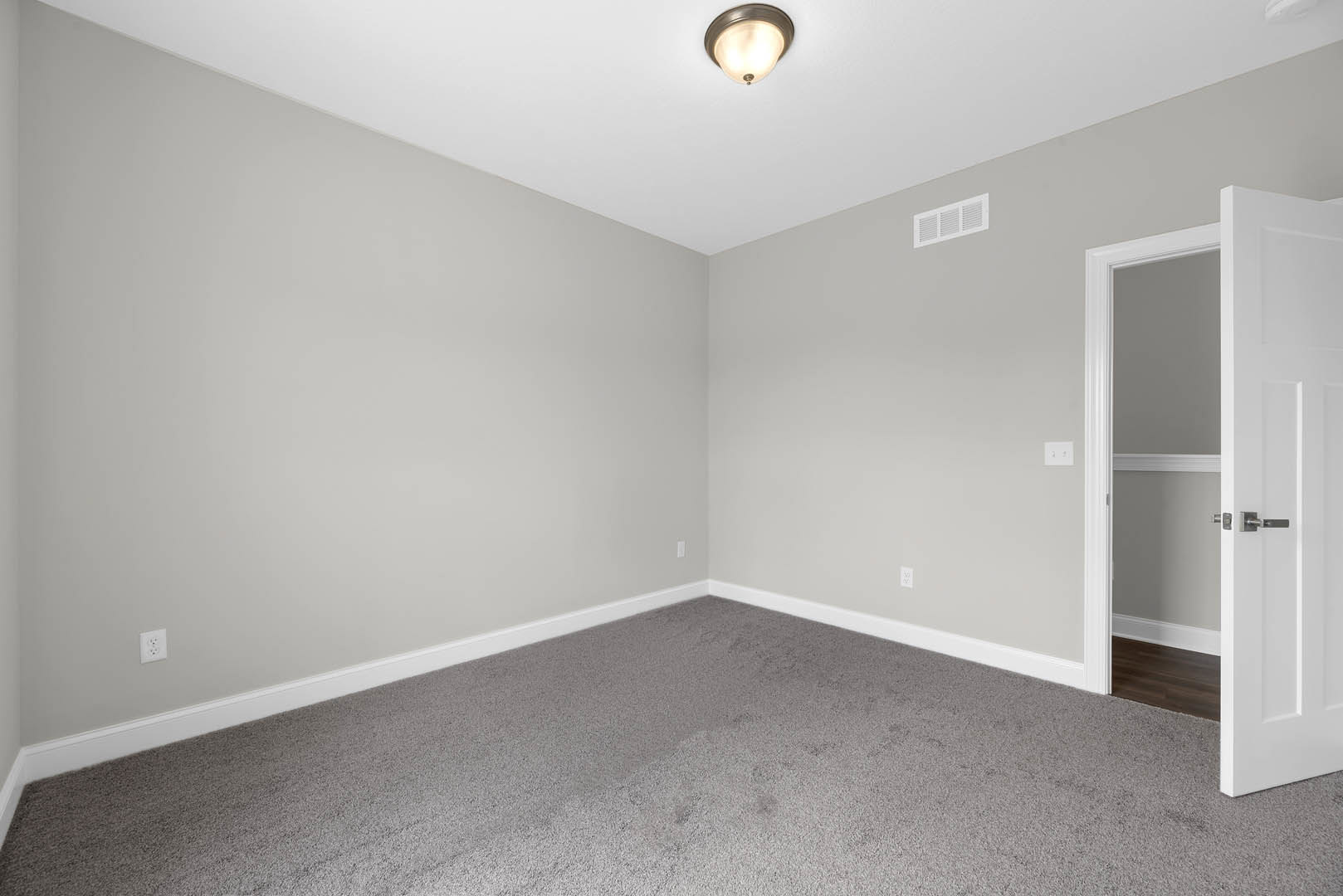 White door with silver handles open to a carpeted room, grey carpet flooring, white ceiling with light fixture and vent, pale walls with molding.