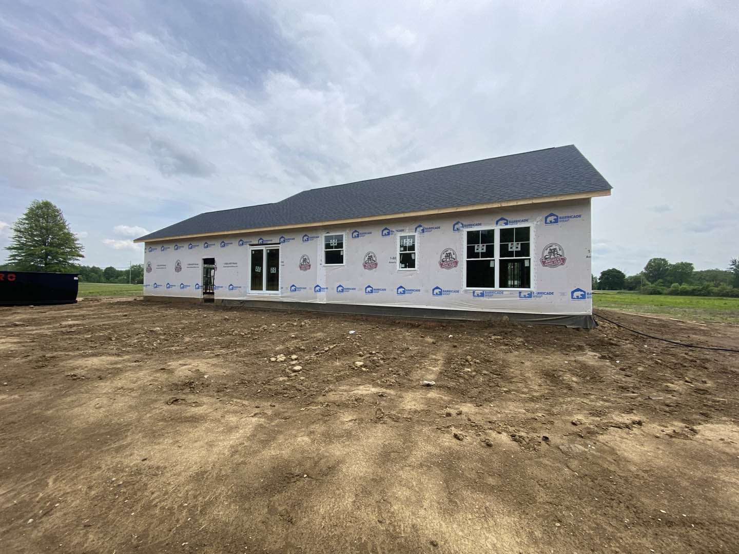 White siding house with blue and white logo panels, dirt lot in foreground, leafy tree to the side, cloudy sky overhead, close-up window visible.